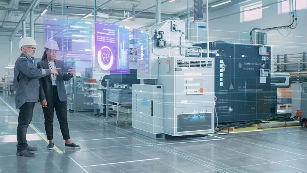 Två människor i säkerhetshjälmar och kostym som står och tittar samt pekar mot industriella maskiner inomhus.
